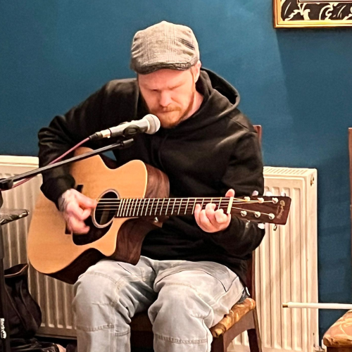 Craig is sitting on a chair, playing an acoustic guitar and singing into a microphone. The backdrop is a blue wall with a framed picture. Craig is indoors, and a guitar case is visible on the floor.